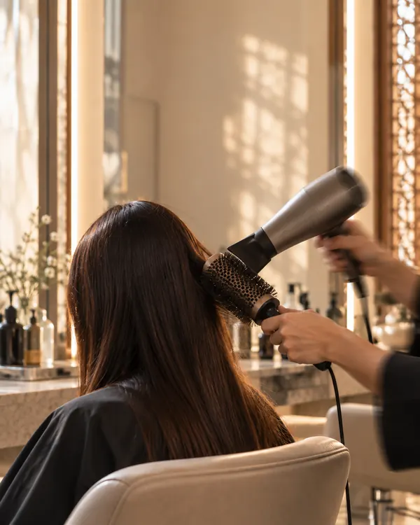 A stylist blow-drying a client's long dark hair with a round brush.